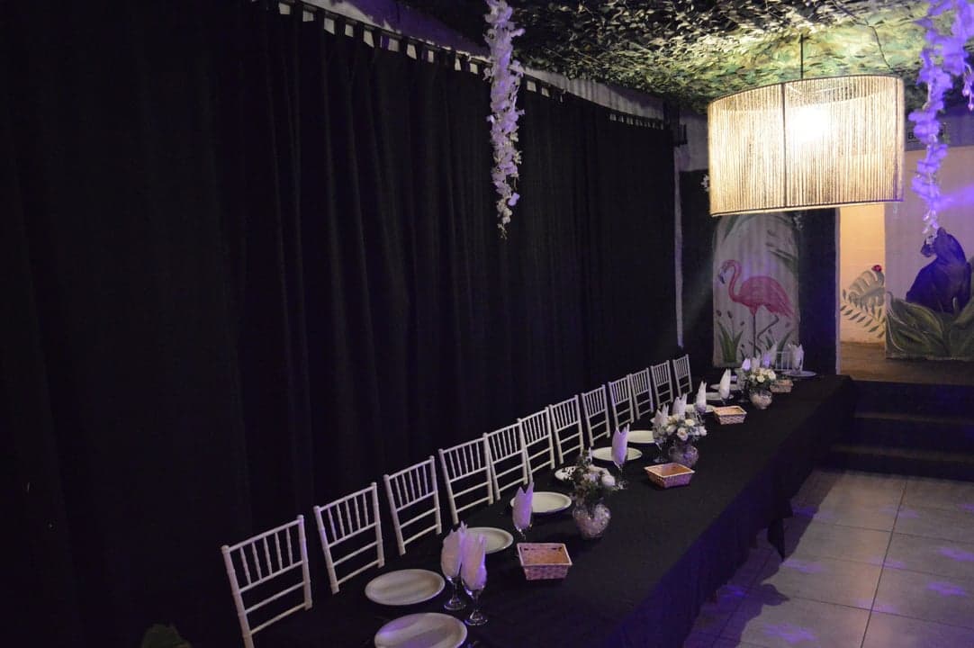 Mesa larga elegante con sillas tiffany blancas, cortinas negras y mural de flamenco